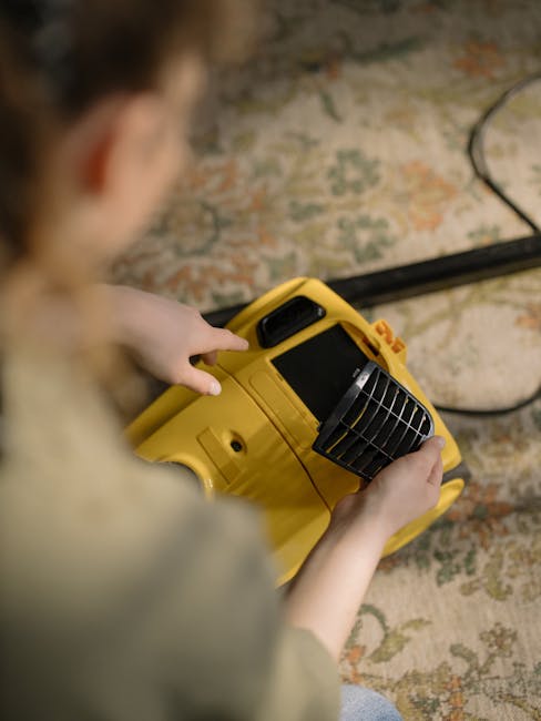 Close-up of a person using a yellow portable fan heater or blower on a patterned carpet, with their hands holding the device and fingers pressing buttons. The carpet features a floral design with mute