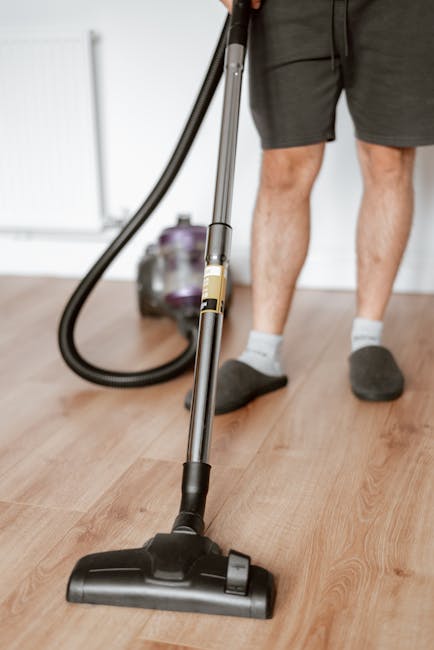 Close-up of a person using a yellow portable fan heater or blower on a patterned carpet, with their hands holding the device and fingers pressing buttons. The carpet features a floral design with mute